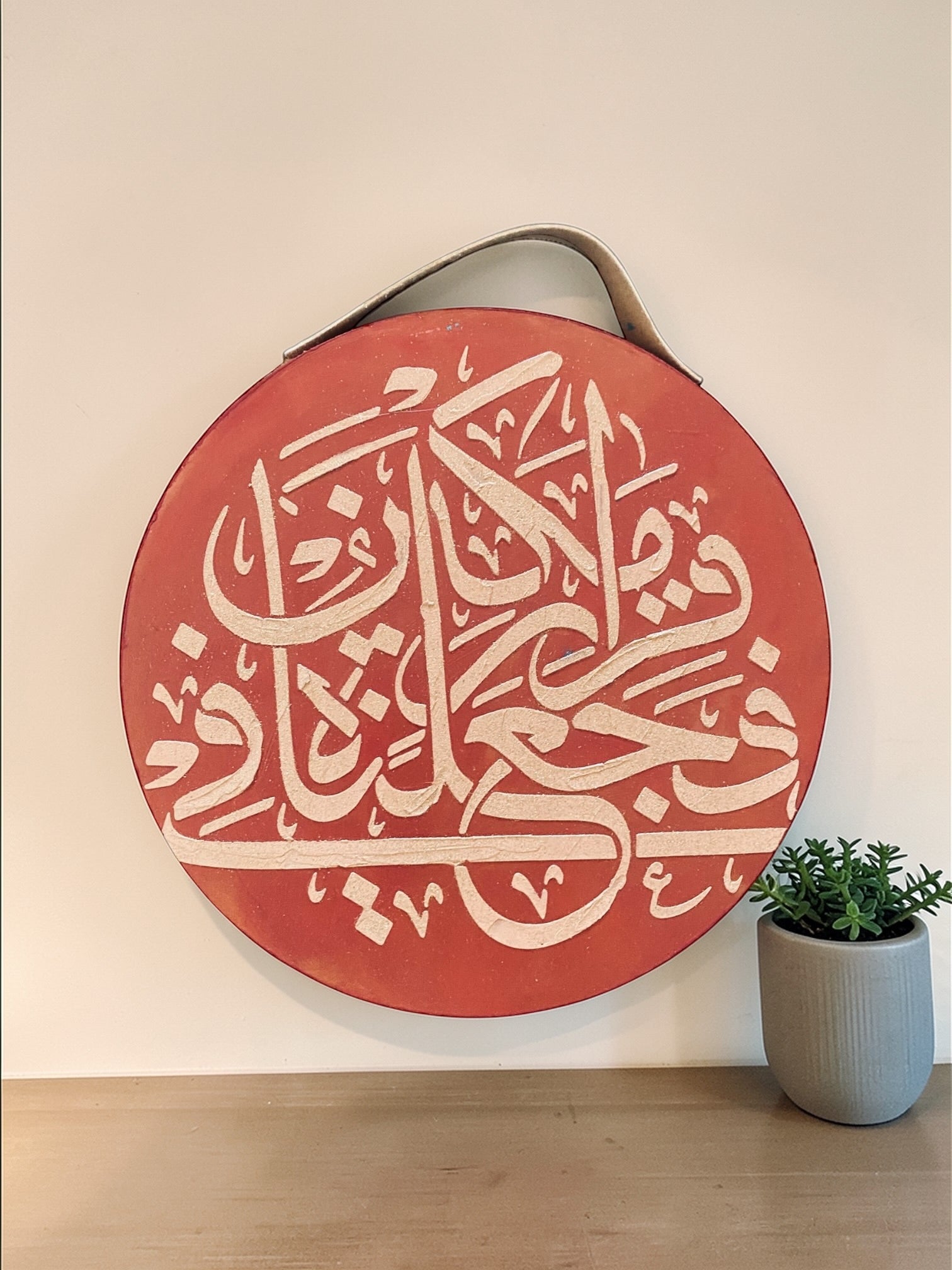 Decorative circular wall art with Arabic calligraphy on a red background, placed on a wooden surface with a plant.