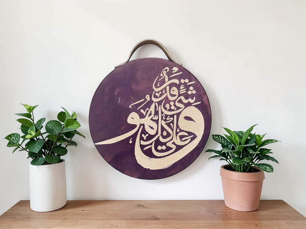 Round decorative piece with Arabic calligraphy on a wooden surface with two potted plants.