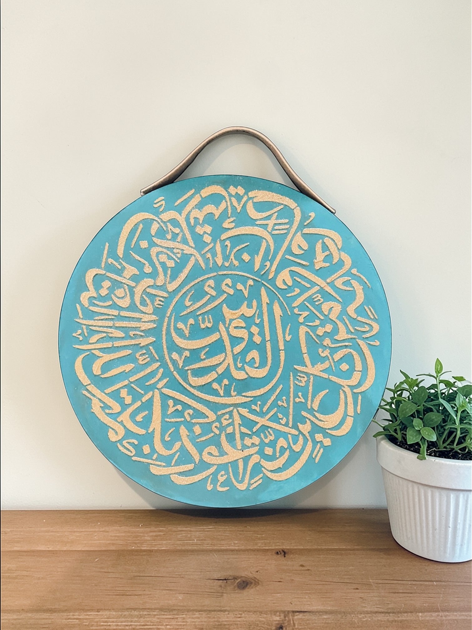 Decorative circular wall art with Arabic calligraphy on a light blue background, mounted on a wooden surface with a plant in the corner.