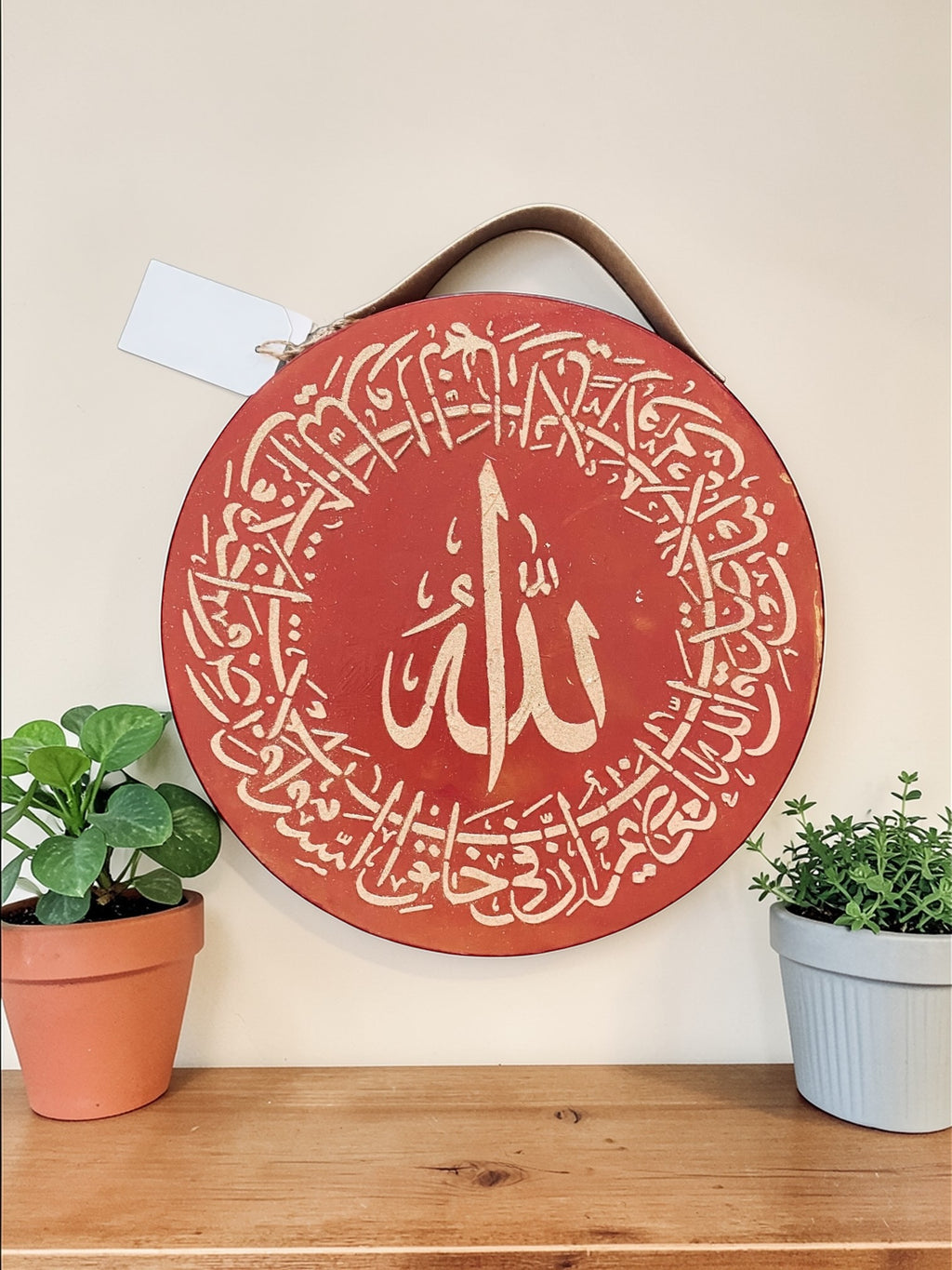 Decorative round sign with Arabic calligraphy on a wooden shelf with plants.