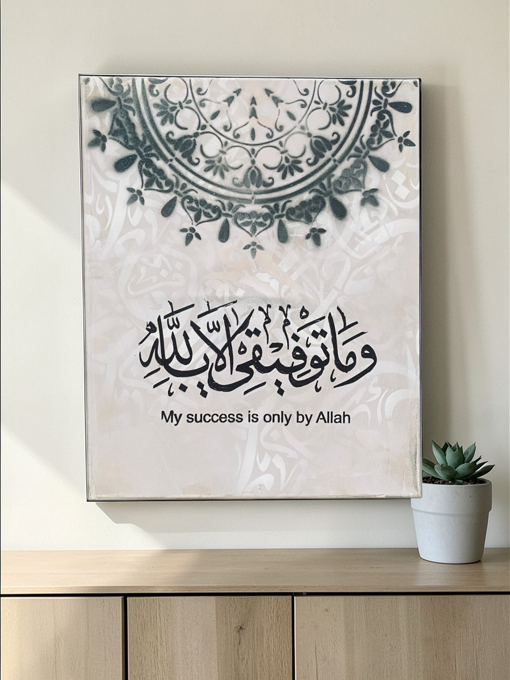 Decorative wall art with Arabic calligraphy and English text on a light background, placed on a wooden surface.
