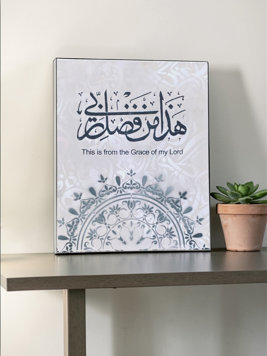 Decorative wall art with Arabic calligraphy and English text on a white background, placed on a wooden surface.