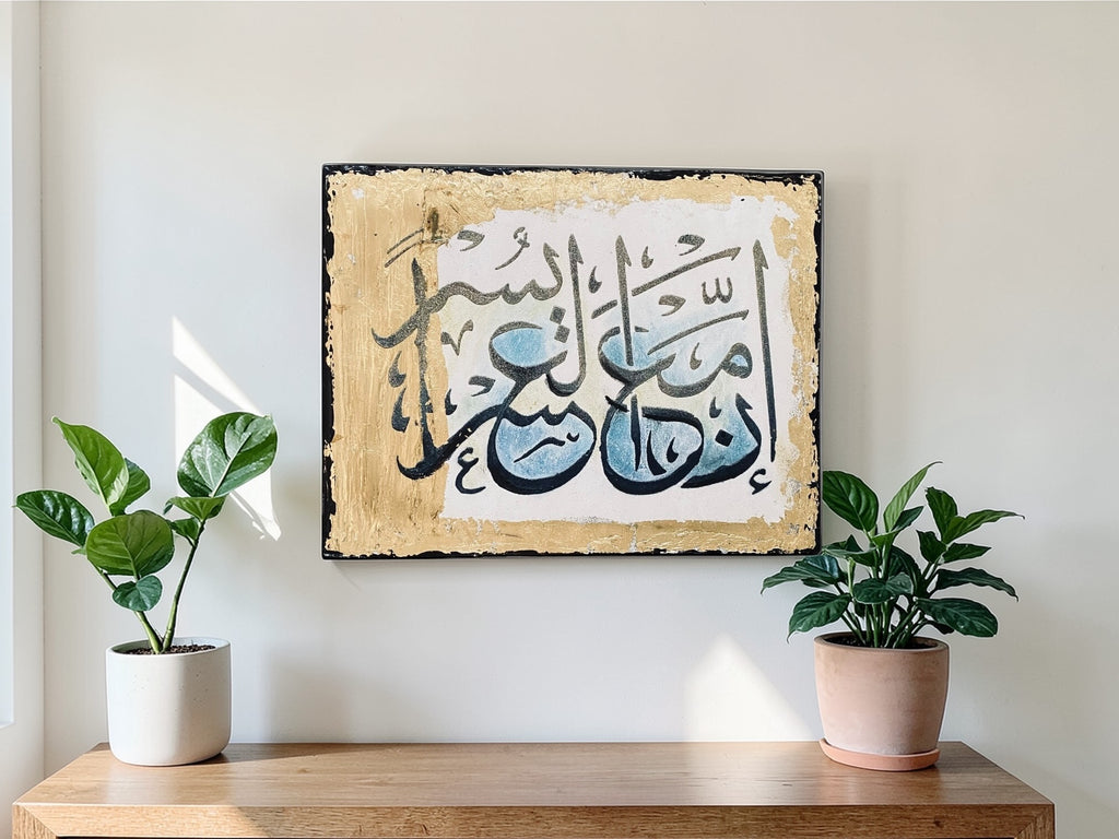 Framed Arabic calligraphy artwork on a wall above a wooden shelf with two potted plants.
